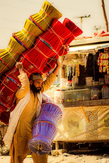 A man with a beard and dressed in traditional clothing is carrying a large bundle of colorful, tightly wrapped paper or fabric objects on his shoulder, with the majority being red, and some purple and patterned. He is walking on a street with small shops and stalls visible in the background, which contain various items including plastic containers, fabrics, and wrapped goods. The scene appears to be outdoors with bright, natural lighting and a clear sky. The man's posture shows he is balancing the weight of the bundles, which rest diagonally across his body. This image relates to waste management through the visual representation of carrying debris or recyclable materials, aligning with independent or private waste handling practices managed by companies like Waste Collection Lambeth, especially in busy market areas where on-site clearance or alternative rubbish removal services are common.