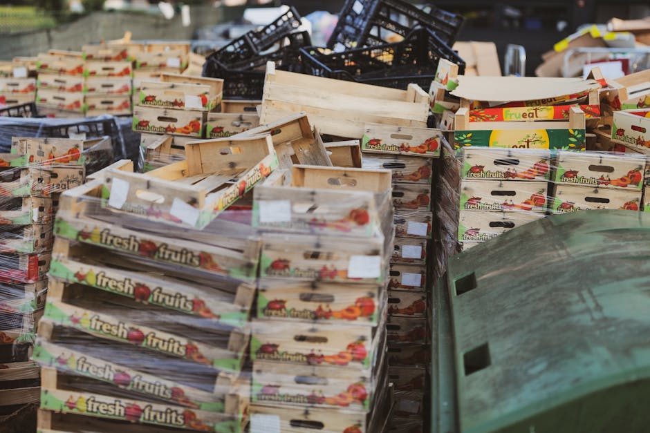 A large collection of empty wooden crates labeled with colorful 'fresh fruits' branding are stacked on a paved surface outdoors, with some crates leaning or piled unevenly. The crates vary in size and condition; some are new, others show slight wear or discoloration, with a natural wood finish. In the background, there are black plastic crates stacked haphazardly, and parts of a green waste bin or container are partially visible in the lower right corner of the image. The scene appears to be in an outdoor setting, possibly a marketplace or storage area, with natural lighting casting soft shadows. The overall impression is of waste or surplus produce packaging ready for disposal or collection, reflecting a typical scenario for independent waste handling and rubbish removal services like those provided by Waste Collection Lambeth.