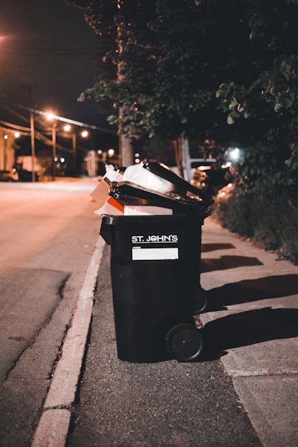 A black wheeled rubbish bin positioned on the edge of a paved sidewalk beside a street at night. The bin is labeled with the text 'ST. JOHN'S' and contains a variety of waste items, including flattened cardboard boxes and plastic containers, some of which are visible overflowing from the top. The scene is illuminated by warm street lighting, casting long shadows on the ground. In the background, there are streetlights, power lines, and out-of-focus trees and parked vehicles, creating an urban environment typical for waste collection or private rubbish removal services. The surrounding area appears residential with foliage and a curb neighboring the bin, indicating a suburban or city street setting, which could relate to independent waste handling or alternative rubbish collection options in the context of waste management services.