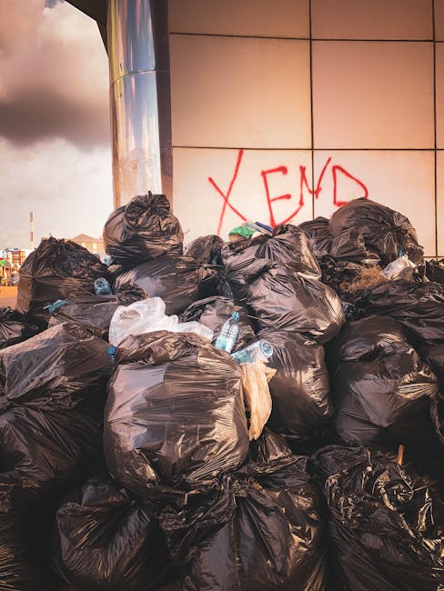 A large pile of black rubbish bags filled with waste, stacked outside an urban building with a tiled exterior wall. Some bags are torn, revealing plastic bottles and other debris inside. The waste bags are positioned on a pavement, with a few scattered items such as plastic bottles and pieces of paper visible among them. In the background, graffiti with the word 'XEND' is spray-painted in red on the building's wall, and the sky is cloudy with a mixture of dark and light tones. The scene suggests an issue with improper waste disposal or collection delays, highlighting the need for regular rubbish removal or alternative waste handling services. Waste Collection Lambeth may be associated with addressing such collection problems, echoing the importance of proper rubbish management in urban environments.