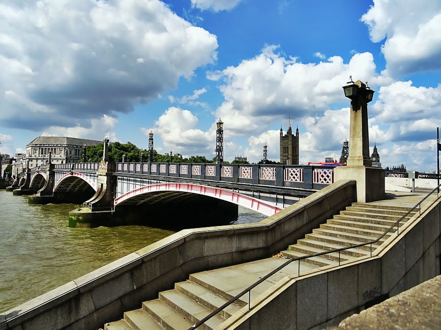 A view of Westminster Bridge crossing the River Thames in London, showing the bridge's stone balustrades painted with pink and white accents, supported by stone arches partially submerged in the water. In the foreground, a concrete staircase with metal handrails leads down to the riverbank, which appears muddy and slightly weathered. On the riverbank, there are stone crenellations and a small section of pavement visible. In the background, Westminster Abbey’s tall, Gothic-style tower with spires rises above the skyline, surrounded by several ornamental metal lampposts mounted on tall, decorative stone pillars that line the bridge. The sky overhead is partly cloudy with large, fluffy clouds and patches of blue. This scene is set in an accessible riverside environment in central London, highlighting the historic architecture and urban landscape that may occasionally require maintenance or waste removal interventions, especially in areas close to busy tourist spots and public spaces, managed by local services such as Waste Collection Lambeth with expertise in waste and rubbish removal services.