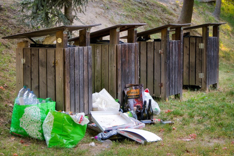 A large pile of black rubbish bags filled with waste, stacked outside an urban building with a tiled exterior wall. Some bags are torn, revealing plastic bottles and other debris inside. The waste bags are positioned on a pavement, with a few scattered items such as plastic bottles and pieces of paper visible among them. In the background, graffiti with the word 'XEND' is spray-painted in red on the building's wall, and the sky is cloudy with a mixture of dark and light tones. The scene suggests an issue with improper waste disposal or collection delays, highlighting the need for regular rubbish removal or alternative waste handling services. Waste Collection Lambeth may be associated with addressing such collection problems, echoing the importance of proper rubbish management in urban environments.