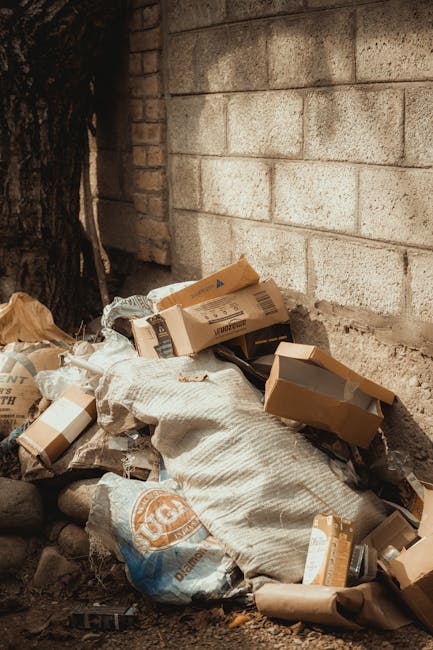 A pile of mixed waste materials, including flattened cardboard boxes, paper wrappers, and a large woven sack, is gathered on the ground next to a brick wall and a tree trunk. The cardboard boxes vary in size, with some partially torn or creased, and are primarily brown with visible printed labels and barcode markings. The woven sack appears to be made of a coarse fabric with a textured surface, and there are additional paper packaging and plastic bags in the mixture. The waste is loosely stacked, with some items leaning against the brick wall, and the scene is set outdoors on a dirt surface, suggesting an informal or private collection point for rubbish removal. The overall environment reflects a typical spot for waste awaiting collection or disposal, consistent with services provided by companies like Waste Collection Lambeth specializing in independent waste handling and rubbish removal for domestic or commercial clients.