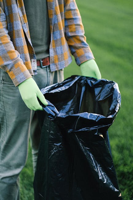 A person wearing a yellow and grey plaid shirt, light grey trousers, and bright green gloves is holding open a large black rubbish bag lined with a silver interior. The person is standing outdoors on a grassy area with a blurred green background, indicating a park or garden setting. The individual appears to be in the process of collecting or disposing of waste, consistent with waste management activities. This scene suggests a private or on-site waste collection scene, possibly related to an independent rubbish removal service such as Waste Collection Lambeth. The black rubbish bag is partially filled and ready for disposal, emphasizing the task of rubbish collection or clearance in a domestic or community environment. The natural outdoor lighting highlights the textures of the bag and the gloves, and the environmental context aligns with alternatives to municipal waste collection, emphasizing the practical aspect of rubbish handling and disposal.