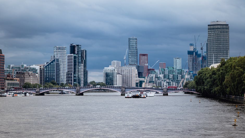 A wide view of a modern urban skyline situated along a river, featuring a mix of high-rise buildings with glass and concrete facades in various shades of white, gray, and dark tones. Several mid-rise and tall skyscrapers dominate the scene, some with distinctive geometric designs, and a few are under construction with cranes visible atop them. The river in the foreground has a calm, slightly rippled surface with a few small boats docked along the water’s edge, and a stone bridge with arch supports spans across the river, connecting two sides of the city. On the right side, there is a row of dense greenery with trees lining the riverbank, providing a natural contrast to the urban environment. The sky overhead appears overcast with thick, dark clouds, suggesting an impending storm or recent rainfall. This scene exemplifies the bustling cityscape typical of a major financial district, with a backdrop that reflects contemporary architecture and urban development, aligning with the context of alternative waste management options available for dense city areas, as managed by Waste Collection Lambeth.