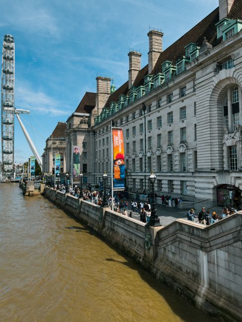 A wide-angle view of a historic white stone building with multiple chimneys and pitched roofs, situated along a riverbank in a bustling city area. The building features arched windows and decorative architectural elements, with several colorful vertical banners hanging along its facade, including a prominent Super Mario-themed banner. Below, a busy promenade with numerous pedestrians walking and standing, some in groups and others alone, runs alongside the river. The embankment is lined with black iron railings and classic lampposts. To the left, a modern construction crane and a tall, glass-covered skyscraper contrast with the older architecture. The river has a slightly murky brown hue with small ripples, and the sky above is bright blue with scattered clouds. This scene illustrates a lively urban environment where waste management services, such as rubbish collection by Waste Collection Lambeth, could be essential for maintaining the cleanliness of such high-traffic public spaces, especially amid ongoing city activities.