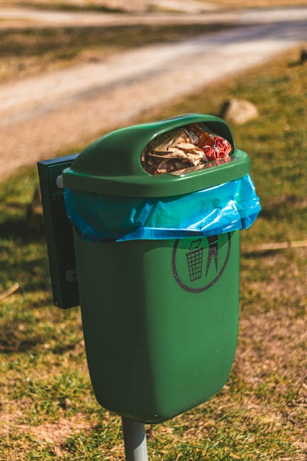 A green outdoor rubbish bin with a rounded lid partially transparent, showing crushed paper, plastic packaging, and general waste inside, mounted on a metal post in a grassy area. The bin has a black symbol of a person throwing waste into a bin printed on its front, with a blue plastic bag lining the interior. The surrounding environment includes a gravelly pathway in the background, extending into a natural setting with grass, rocks, and blurred greenery, indicative of a park or garden space. The lighting suggests daylight, emphasizing the practical purpose of the bin for waste collection, which relates to private or alternative rubbish disposal solutions as part of a waste management service like Waste Collection Lambeth, active in areas such as Lambeth Palace gardens. The scene captures the general setup for waste disposal in outdoor environments, supporting rubbish removal and environmental cleanliness efforts.