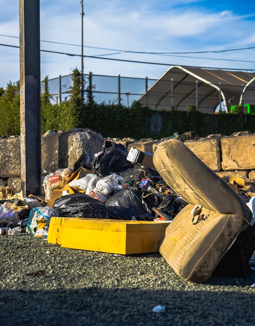 A roadside scene featuring a pile of mixed rubbish including black plastic garbage bags, a yellow plastic container, a worn beige car tyre, and various smaller waste items, all situated on a gravel surface adjacent to a low stone wall made of rough, irregular stones. Behind the wall, there is a green hedge and some trees, with a sports field or tennis court enclosed by a black fence in the background. Utility poles and overhead wires are visible against a partly cloudy sky, indicating an outdoor setting. The sunlight casts shadows, highlighting the textures of the discarded waste materials and the surrounding environment. The scene illustrates unmanaged rubbish accumulation, which waste collection services like Waste Collection Lambeth might be called to clear, especially where private or independent disposal options are used to handle waste outside of regular council collections.