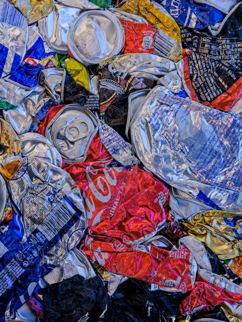 A close-up view of a pile of crushed and crumpled aluminum cans, predominantly silver with scattered red, blue, yellow, and black labels and branding. The cans vary in size and are made of lightweight, reflective aluminum material, with some showing visible pull-tabs and embossed details. They are stacked loosely on top of each other, with some overlapping and leaning against one another, creating a textured, chaotic arrangement. The background appears to be part of a waste collection or disposal area, with no identifiable environment beyond the focus on the compressed cans. The lighting highlights the metallic sheen and various colors on the labels, emphasizing the texture and condition of the discarded beverage containers. This scene illustrates the type of waste handled by private rubbish collection services like Waste Collection Lambeth, reflecting on the environmental aspect of aluminum can recycling and disposal in urban waste management.