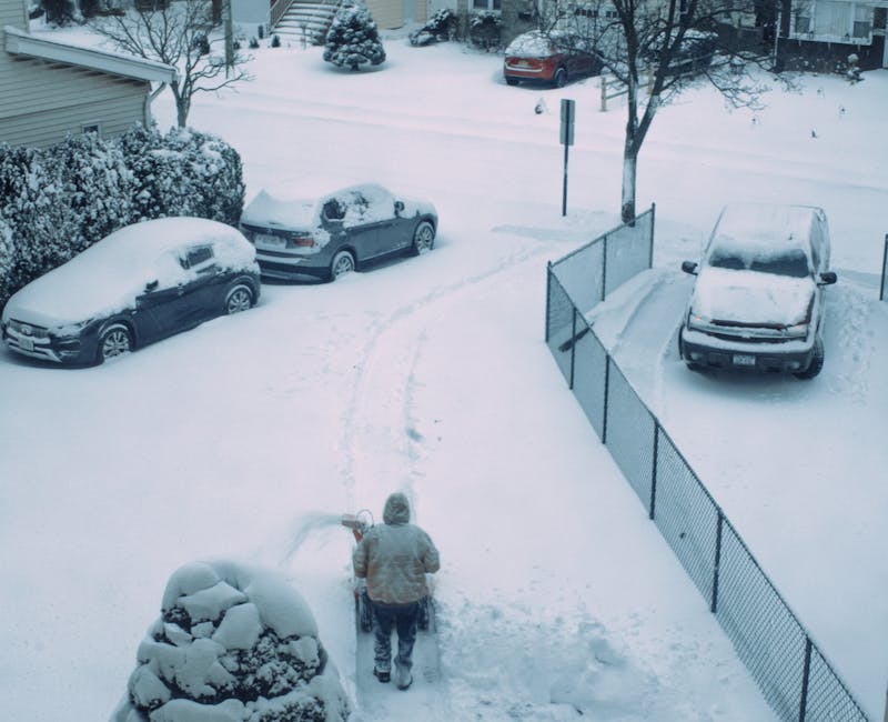 A person dressed in a grey winter jacket and dark trousers is seen from above, walking through a snow-covered residential driveway while clearing snow with a handheld snow shovel. The driveway is surrounded by parked cars, all covered with a thick layer of snow, including a dark-colored hatchback on the left, a silver SUV in the middle, and another vehicle on the right, partially obscured by a black metal fence. The fence runs along the right side of the driveway, separating it from a garden area with snow-covered trees and shrubs. Footprints and snow-clearing tracks are visible on the snowy surface, indicating recent efforts to clear the pathways. In the background, additional snow-covered parked cars and houses are visible, with snow blanketing the entire scene, creating a winter landscape that reflects typical conditions for private property maintenance and rubbish removal in snowy weather. The overall scene depicts an outdoor residential setting with an emphasis on snow clearance amidst winter conditions, associated with independent household waste management routines.