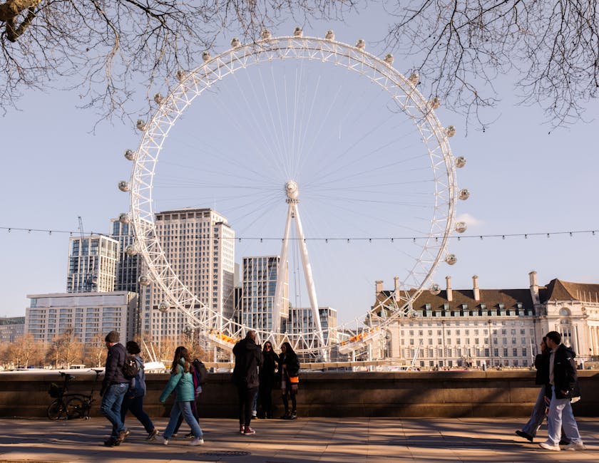 A large observation wheel with a white metal frame and numerous passenger cabins encircling its circumference is prominently positioned against a backdrop of modern high-rise buildings and older architectural structures. The wheel is situated outdoors on a paved area with a stone barrier in the foreground, where several people are walking and standing. The pedestrians, dressed in casual clothing, include individuals and groups, some engaged in conversation or looking up at the wheel. A bicycle leans against the barrier, adding to the urban scene. Behind the wheel, leafless tree branches extend into the sky, which is clear and bright with a few scattered clouds, suggesting an early spring or late autumn day. The overall scene is well-lit with natural daylight, highlighting the textures of the metal structure, clothing, and surrounding architecture. This image subtly relates to alternative waste handling, as it captures an outdoor public space where rubbish removal services might be necessary for events or maintenance, akin to private clearance or onsite cleanup obligations that Waste Collection Lambeth could support.